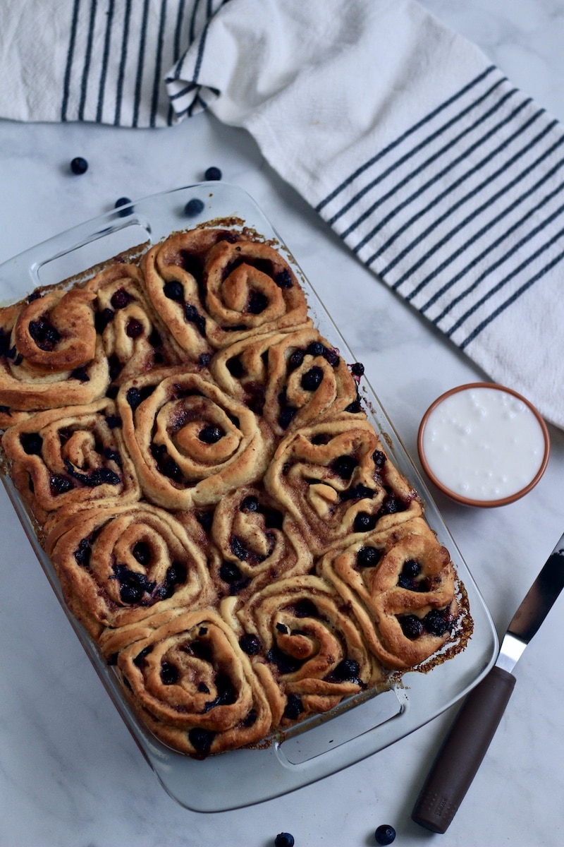Baked lemon blueberry sweet rolls with a small bowl of lemon glaze to the right and an offset spatula to spread the glaze with on the rolls.