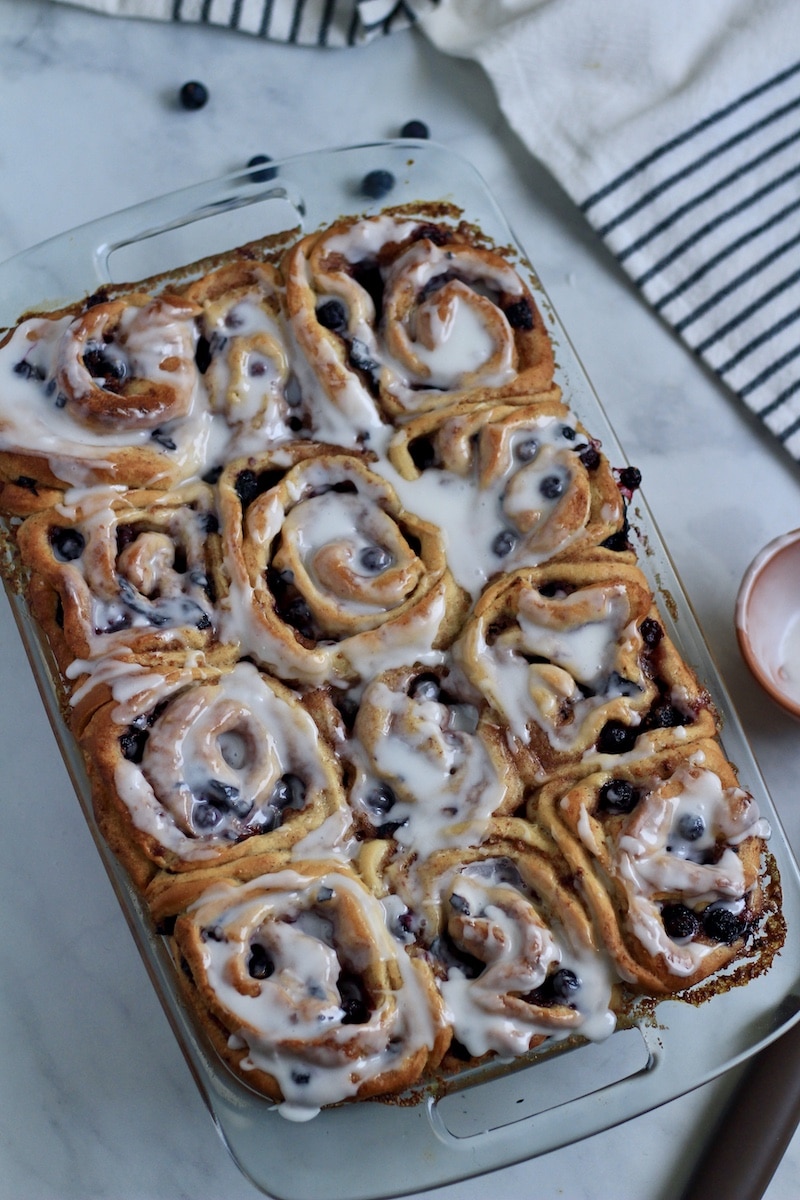 Glazed lemon blueberry sweet rolls in a glass baking dish with a blue and white striped towel in the top right.