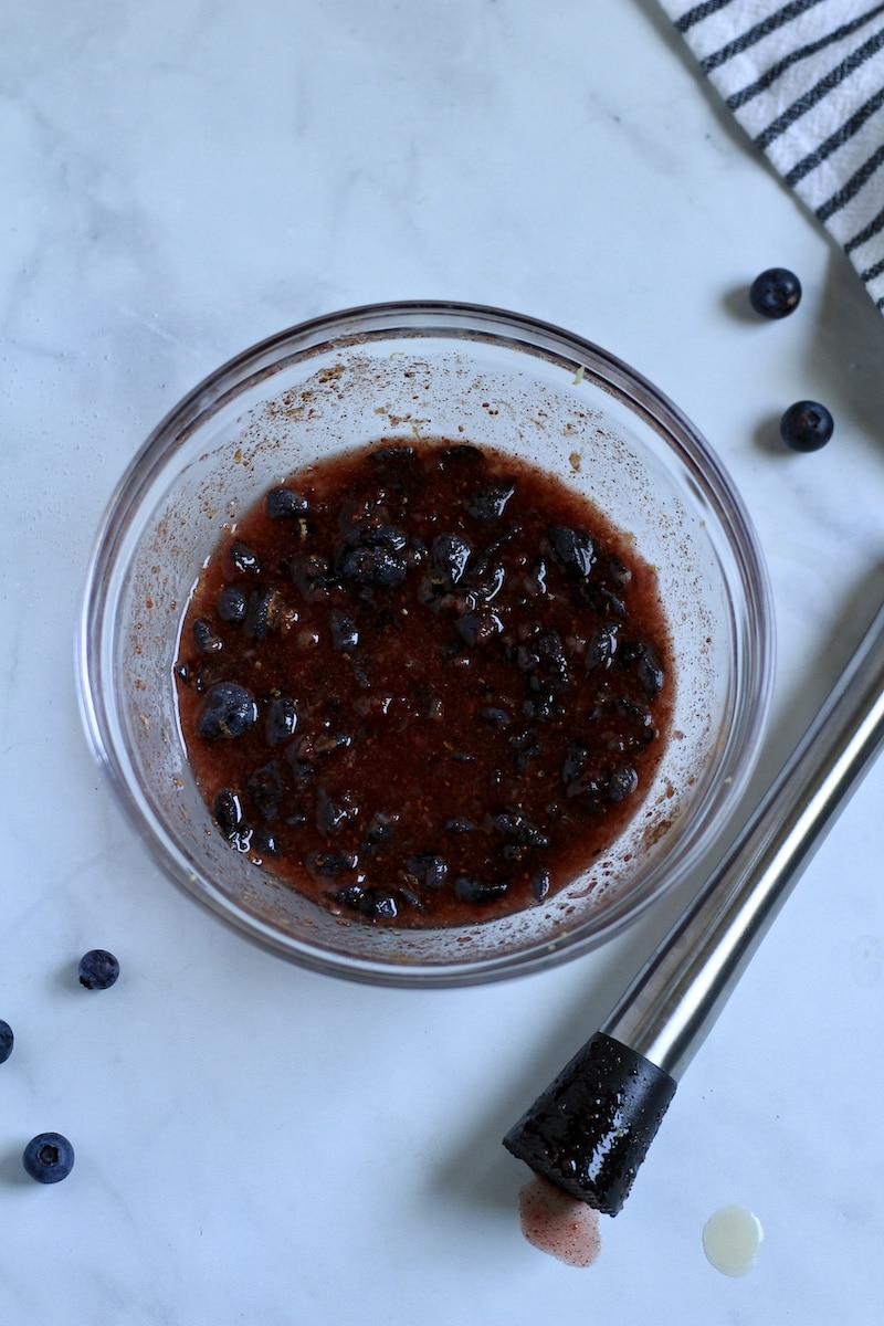 A glass bowl with muddled blueberry filling next to a muddler on a counter.
