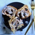 A square photo of a blue plate with three lemon blueberry rolls next to two gold forks.