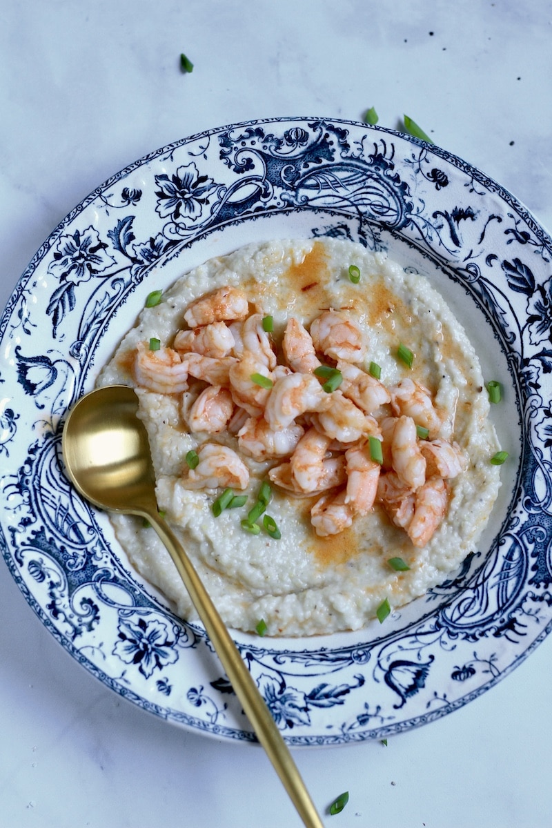 A blue rimmed bowl with chili garlic shrimp and grits and a gold spoon in the bottom left corner of the bowl.