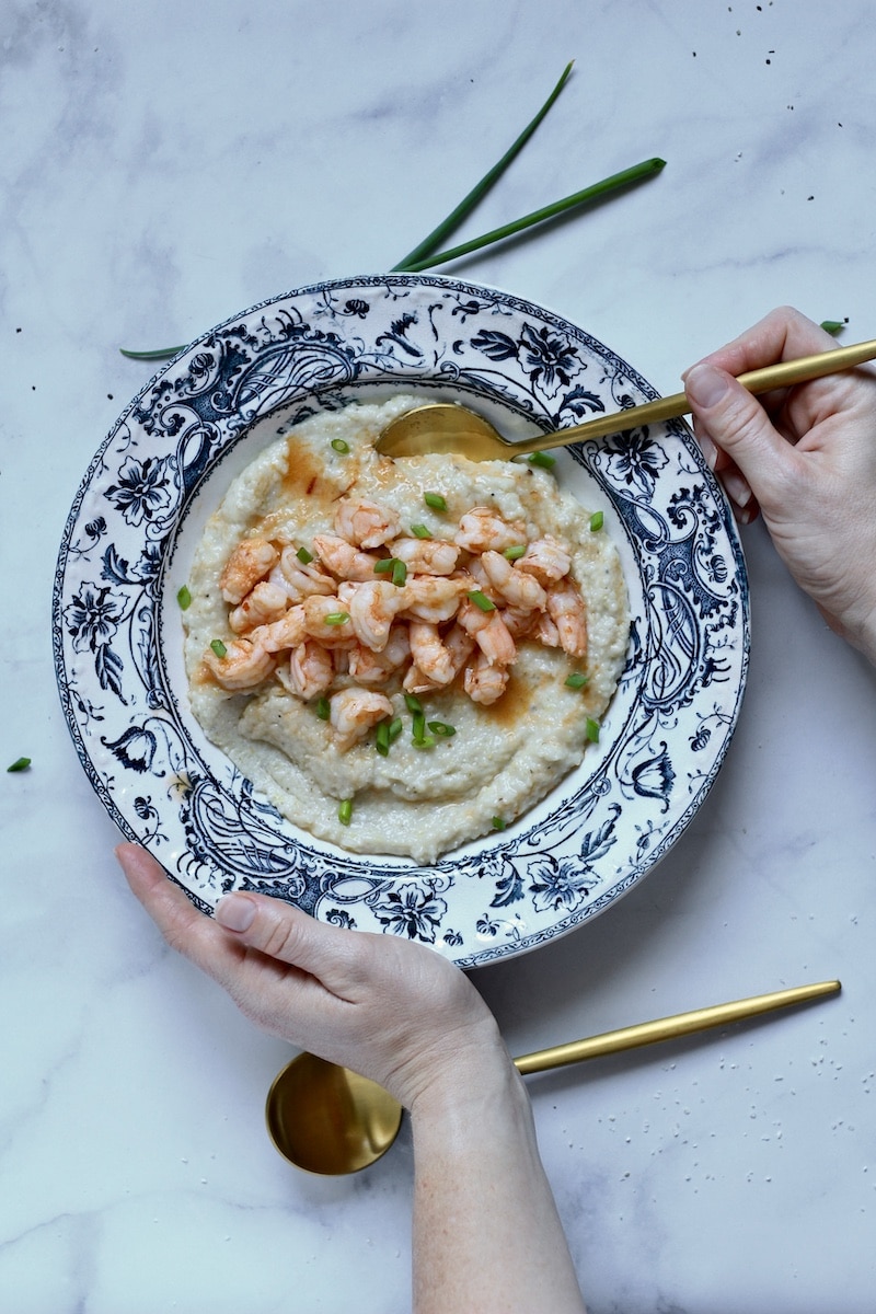 Two hands holding a blue rimmed bowl filled with chili garlic shrimp and garlic butter grits with green onion on top.