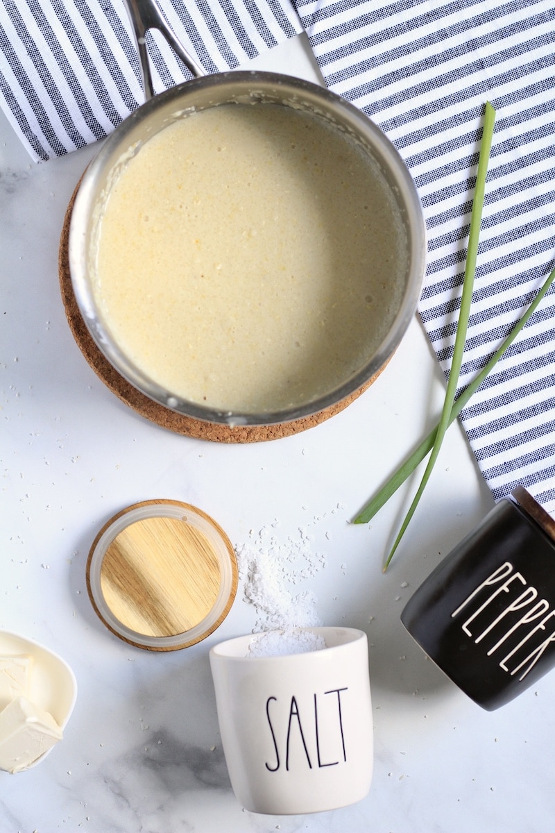 Plain grits in a saucepan before adding garlic butter and spices with a salt and pepper container on the counter.