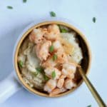 A small orange rimmed bowl with a gold spoon in the right side of the bowl with garlic butter grits topped with chili garlic shrimp.