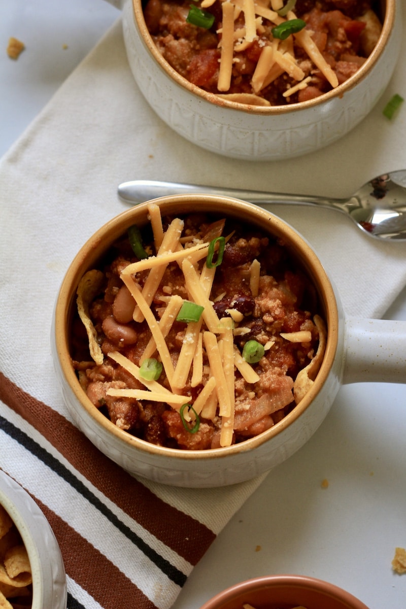 Top down photo or a bowl with a handle on the right with chili topped with cheese and green onion with a spoon above the bowl.