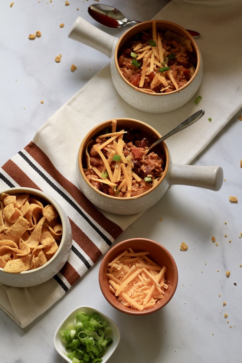 A top down photo of two bowls with handles of frito pie topped with cheese and green onion with bowls of toppings in the front.