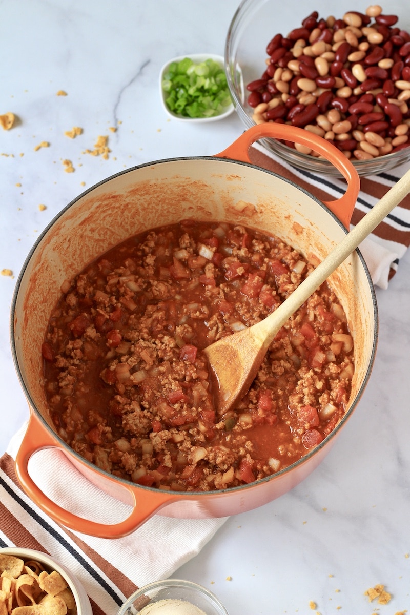 A large orange pot of meat and seasoning before beans get added in with a wooden spoon in the right side of the pot.