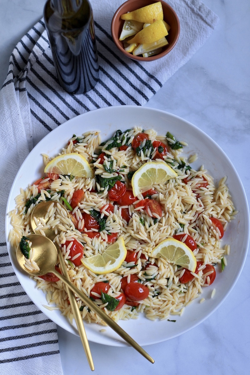 A bowl of lemon orzo pasta salad with lemon wedges and two gold spoons on the left.