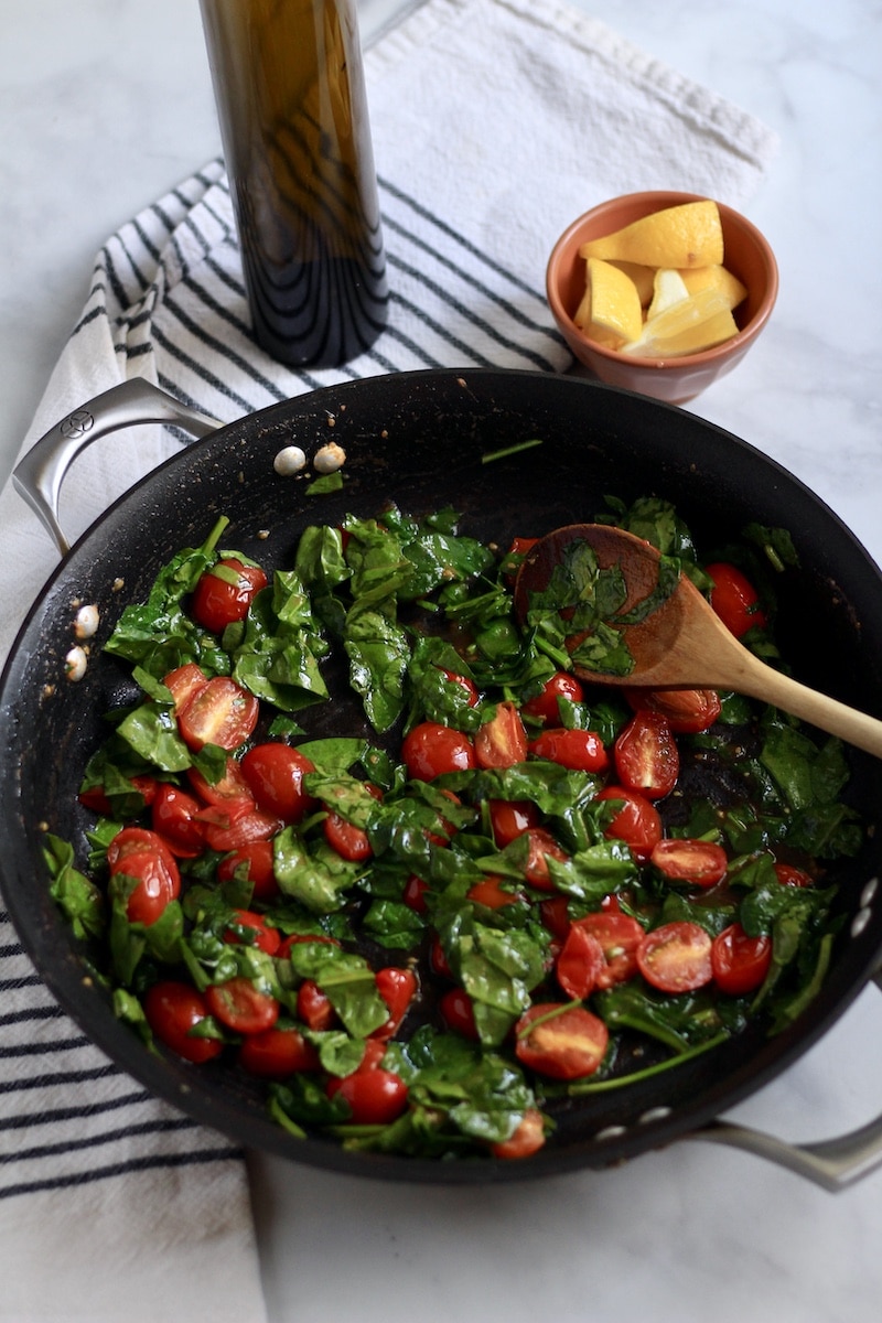 Roasted grape tomatoes in a skillet with chopped spinach and a wooden spoon.