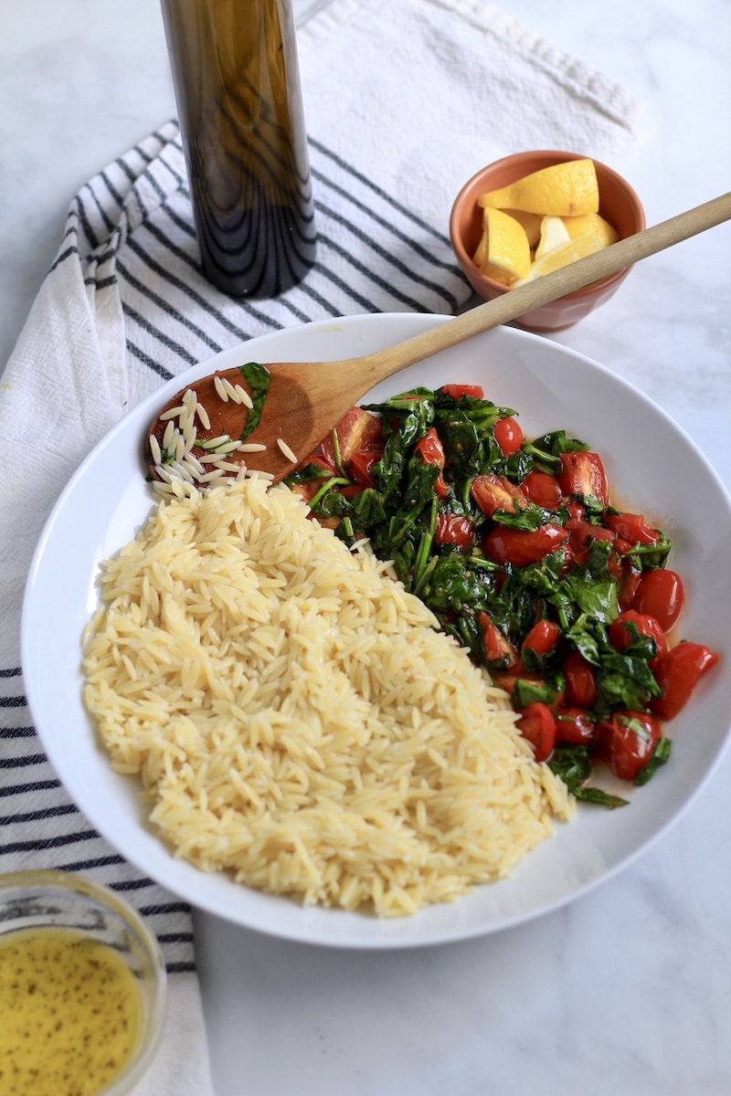 A white bowl with cooked orzo pasta on the left and the cooked spinach and grape tomatoes on the right with a wooden spoon at the top.