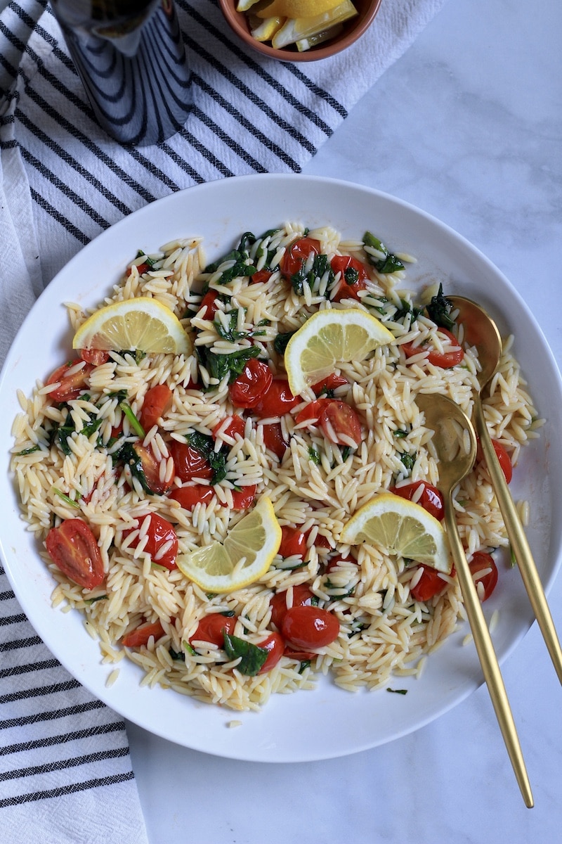 A white bowl with vegan pasta salad and two gold spoons on the right.