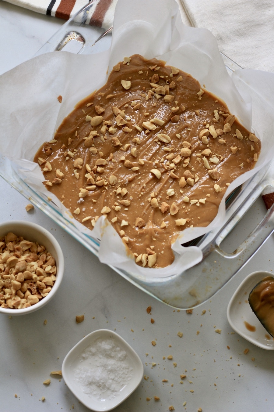 Peanut butter fudge topped with peanuts in a parchment paper lined baking dish with peanuts and salt below.