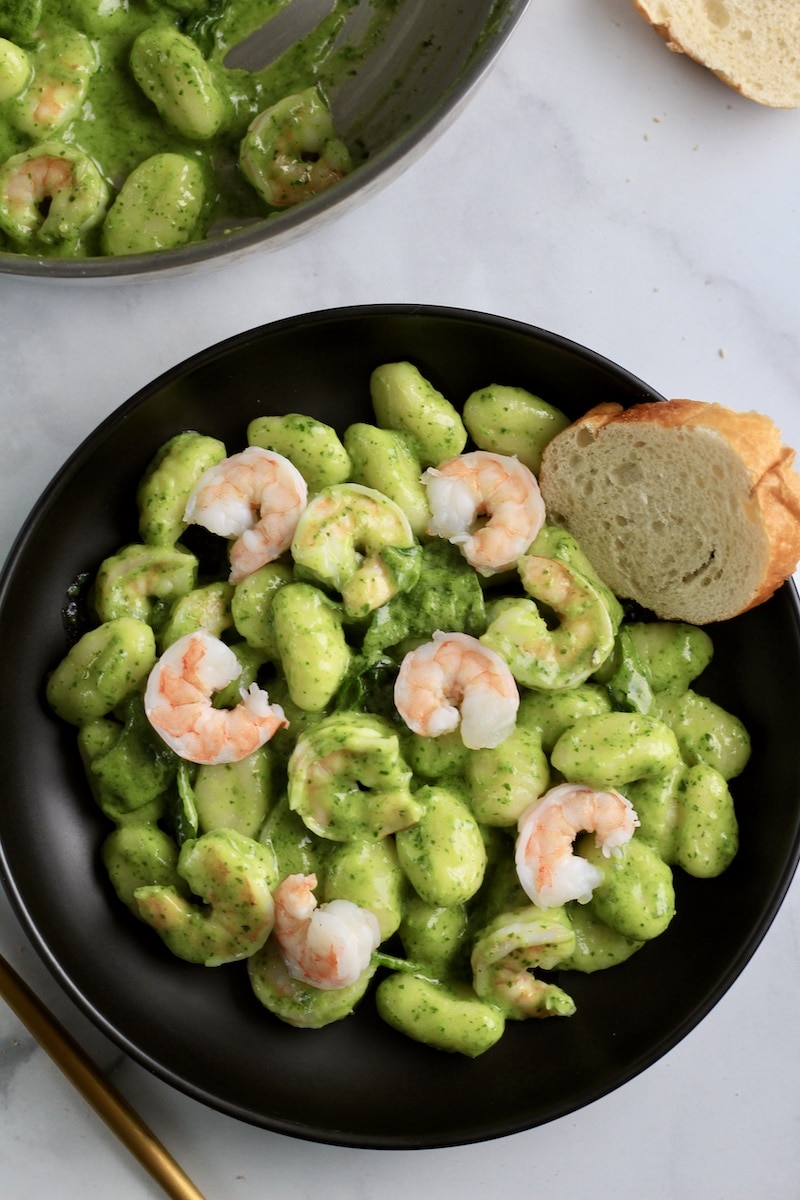 A top down photo of creamy pesto gnocchi with shrimp on a black plate with a slice of bread.