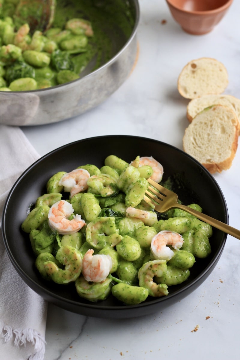 A black plate with creamy pesto gnocchi with shrimp and a gold fork on the right side with slices of bread in the top right corner.