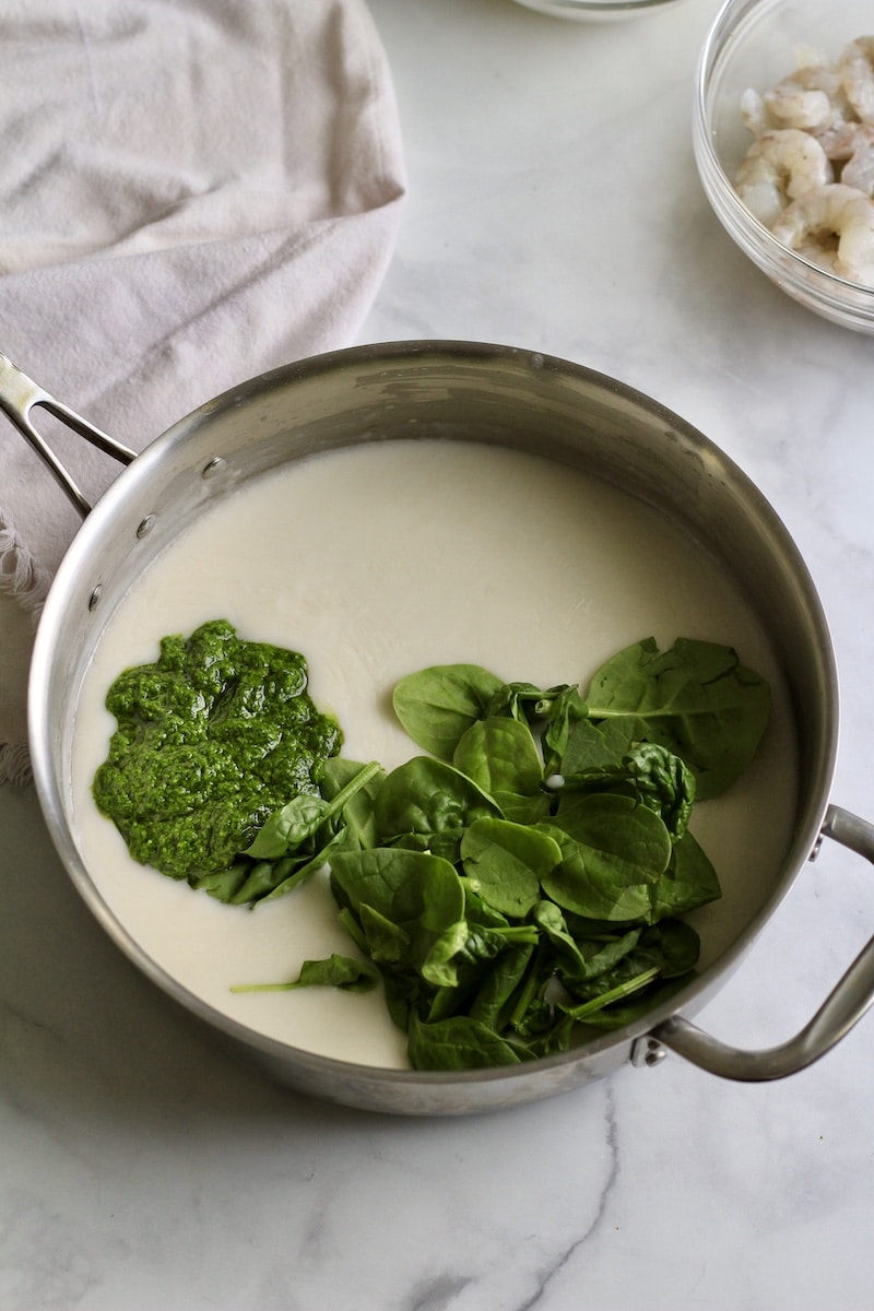 A silver skillet on a white counter with cream sauce, spinach, and pesto before stirring together.