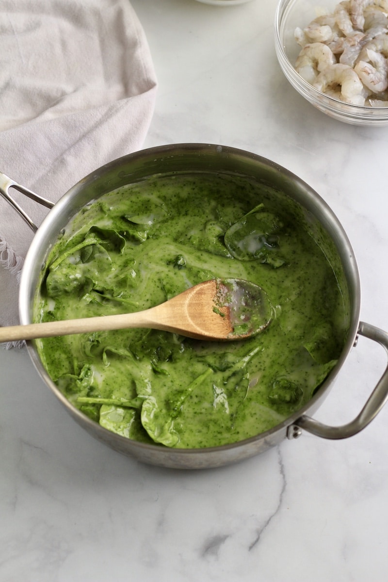 A silver skillet with creamy pesto sauce after stirring with a wooden spoon in the skillet.