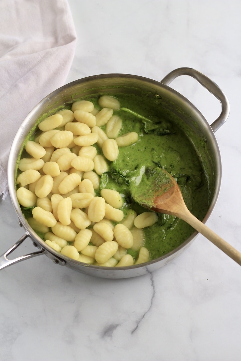 A silver skillet with creamy pesto sauce and gnocchi with a wooden spoon before stirring.