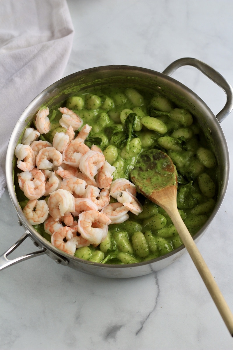 Creamy pesto gnocchi with shrimp and a wooden spoon in a skillet before stirring.