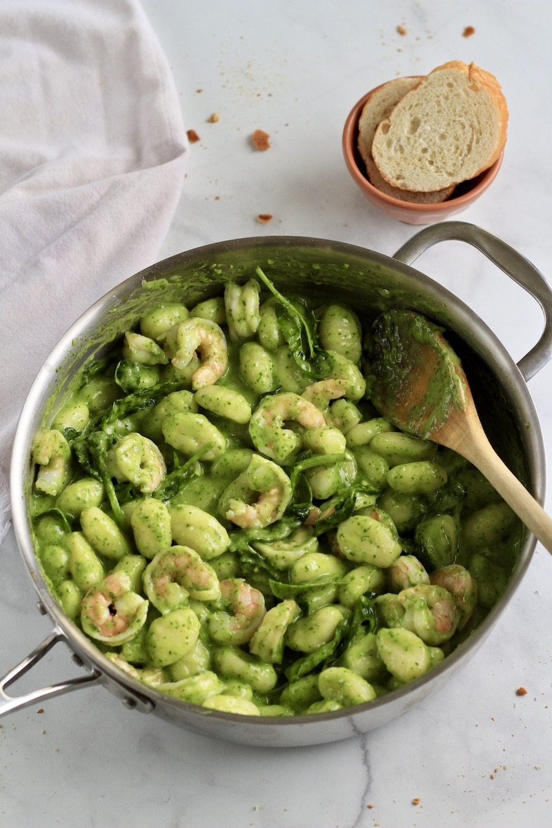 Creamy pesto gnocchi with shrimp in a silver skillet with a wooden spoon and some bread above the skillet.