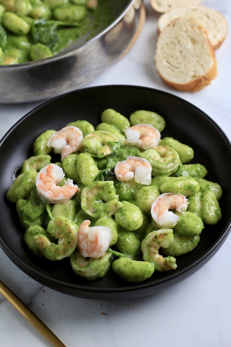 A black plate with creamy pesto gnocchi with shrimp and some bread in the top right of the photo.