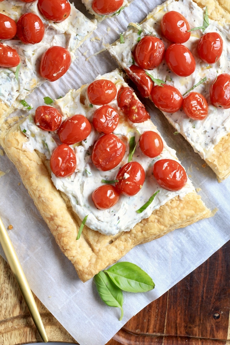 A close up of a slice of roasted tomato tart on parchment paper with basil at the bottom and a gold knife to the right.
