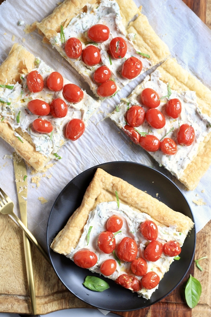 A parchment paper with three pieces of roasted tomato tart with a blue plate in front with another slice of tart and a knife and fork to the left.