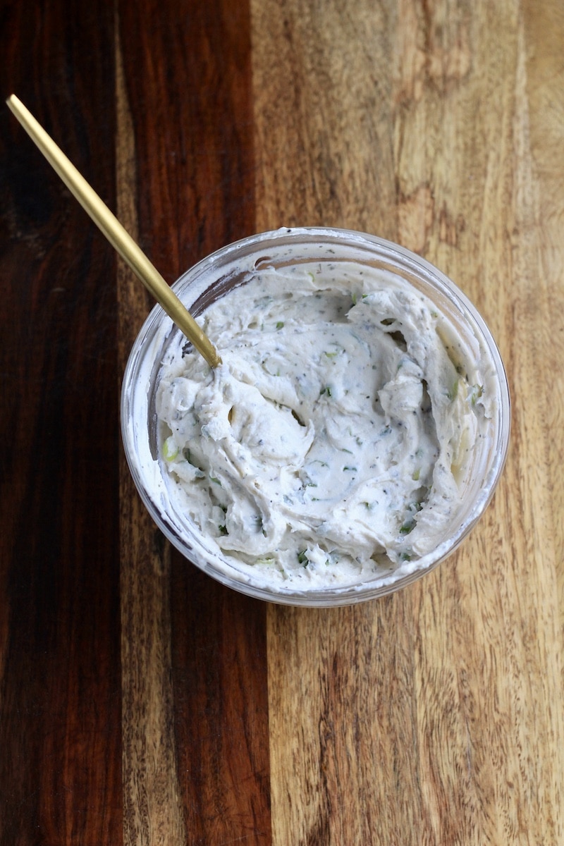 Herbed cream cheese spread in a glass bowl with a gold spoon on a wooden counter.