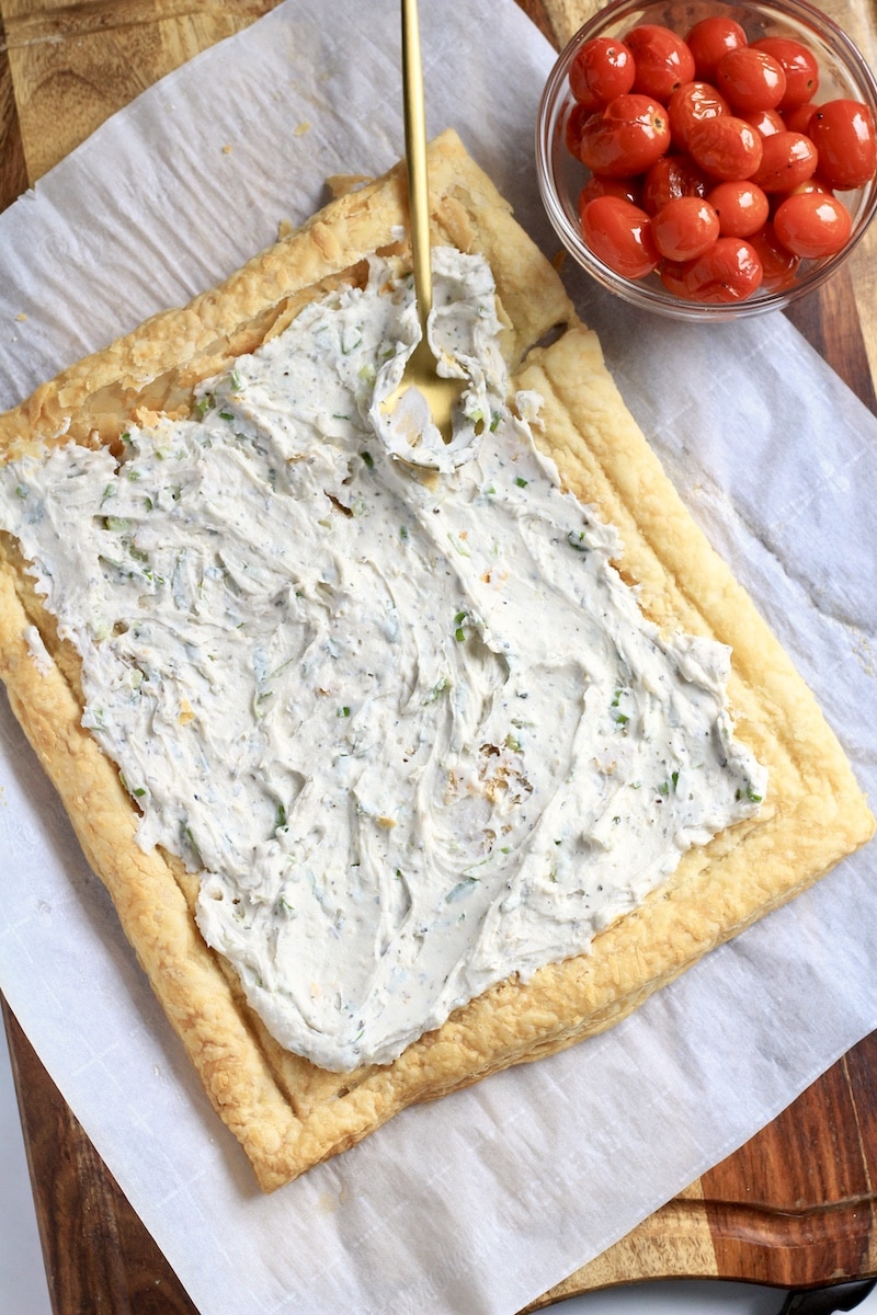 A puff pastry topped with herbed cream cheese with a gold spoon spreading the herbed cream cheese.