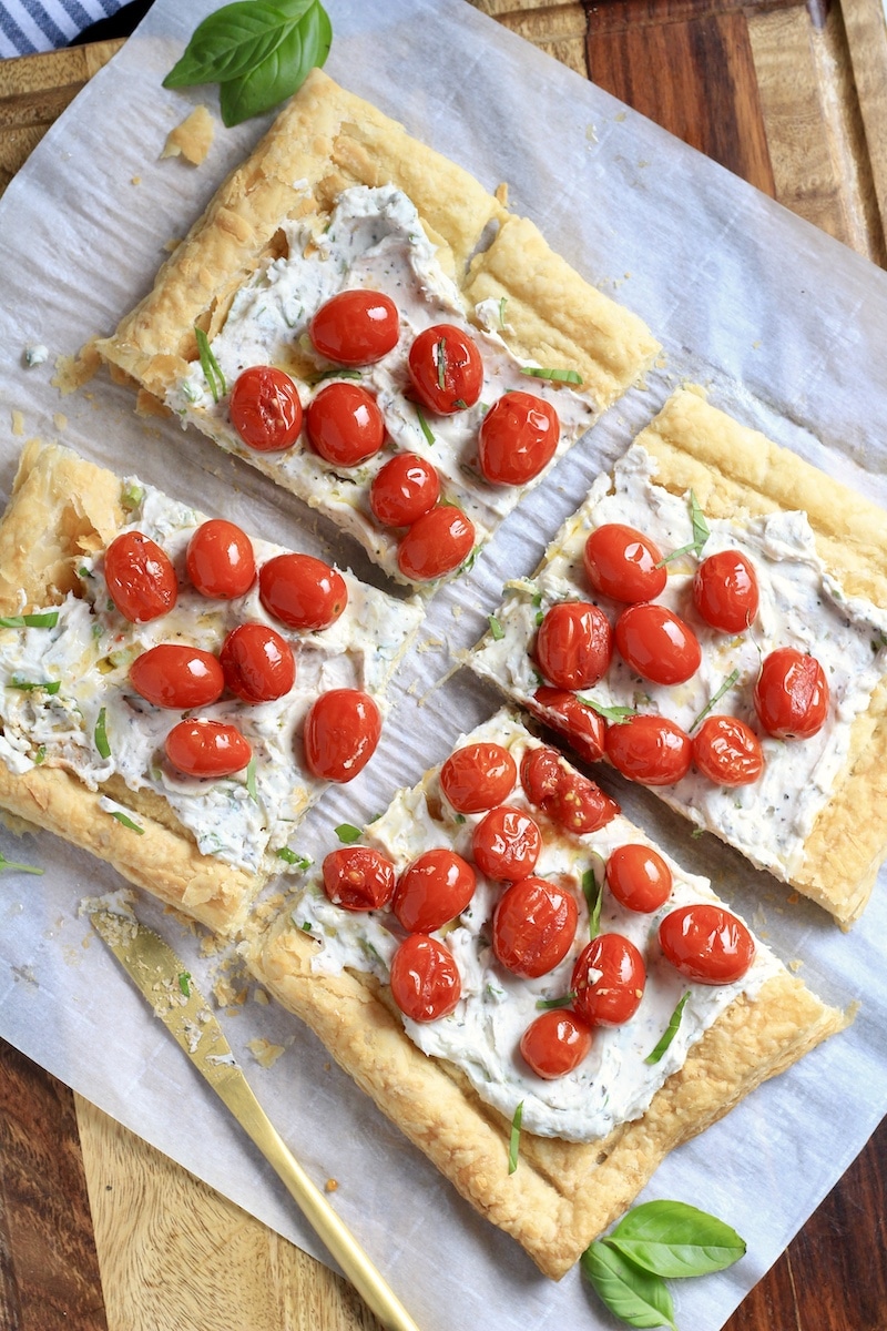 A roasted tomato tart cut into four squares with basil scattered around and a gold knife to the left.