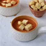 A white bowl with an orange rim filled with tomato and lentil soup and topped with homemade croutons with a small bowl of croutons in the back.