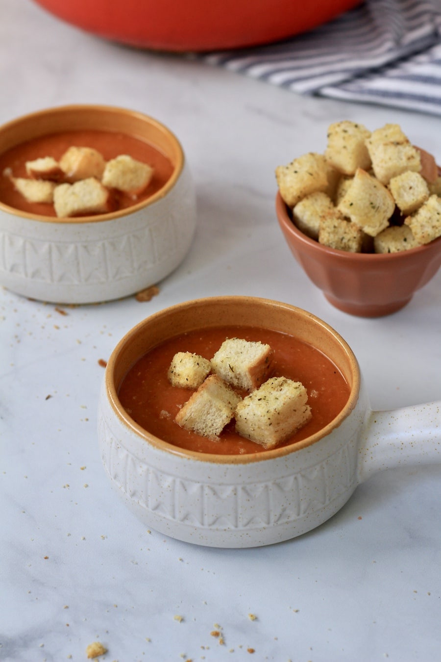 Two small orange rimmed bowls with tomato lentil soup topped with croutons and a small bowl of croutons in the back.