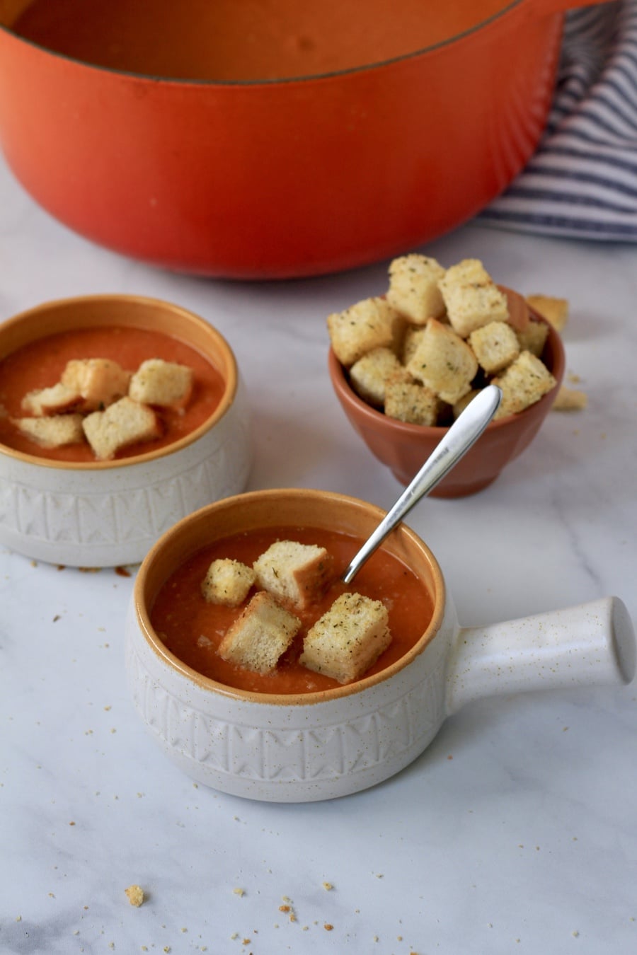 Two small bowls with handles filled with tomato lentil soup and topped with homemade croutons with a small bowl of croutons in the back and a large pot of tomato lentil soup.