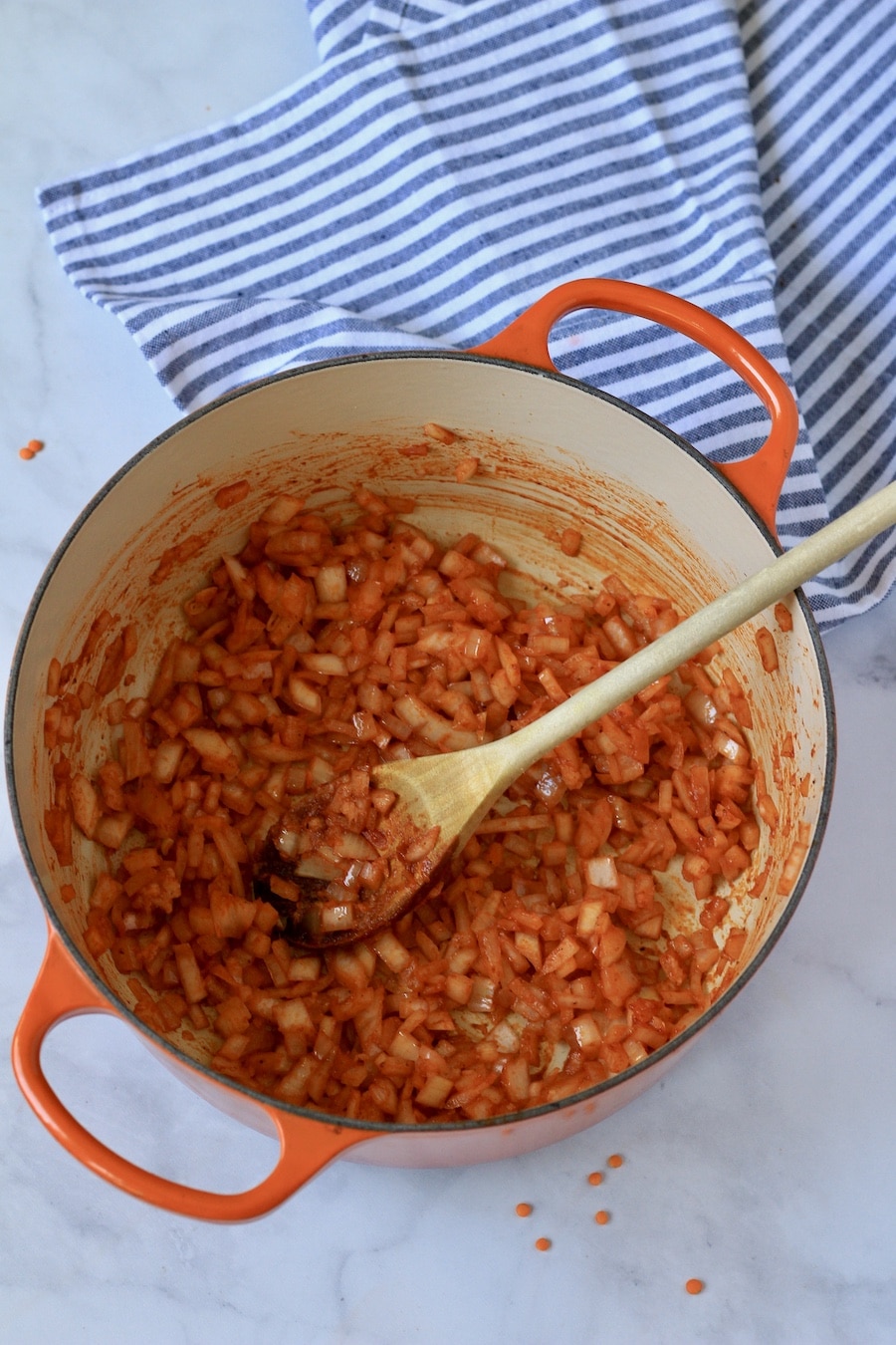 An orange handled dutch oven with tomato paste, garlic, and onions cooking with a wooden spoon.