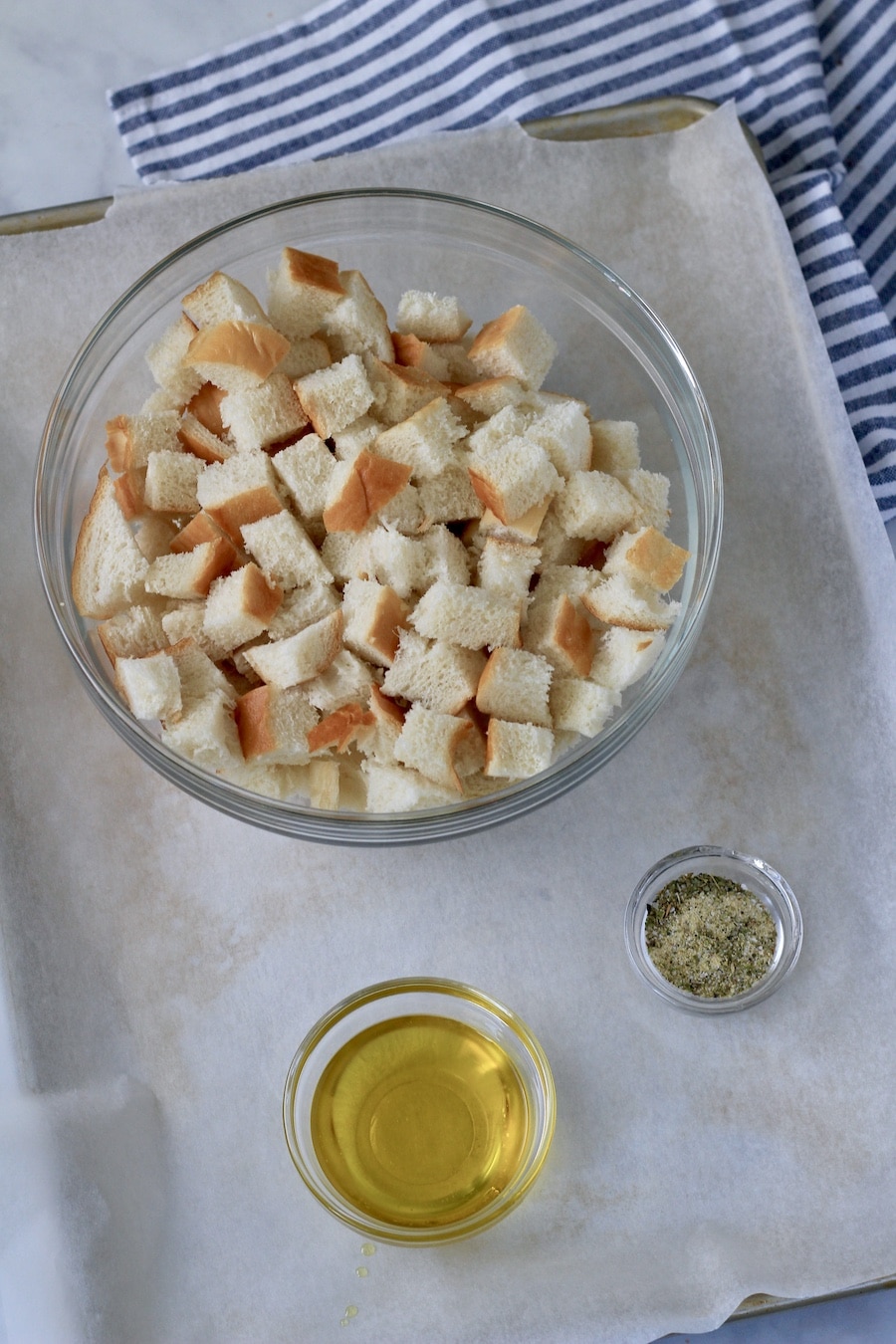 A parchment paper lined baking sheet with cubed bread in a bowl, olive oil in another bowl, and a small bowl of spices for the croutons.