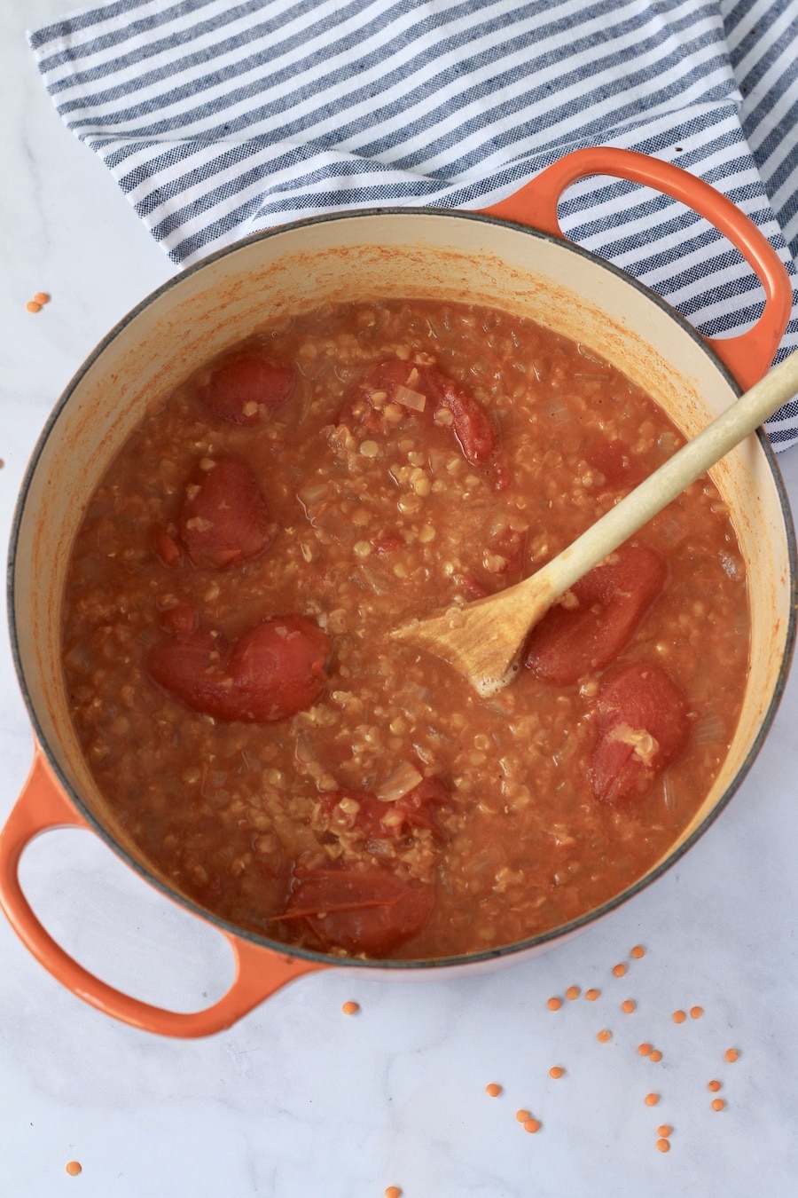 An orange dutch oven with tomato lentil soup after simmering before blending and a wooden spoon in the pot.