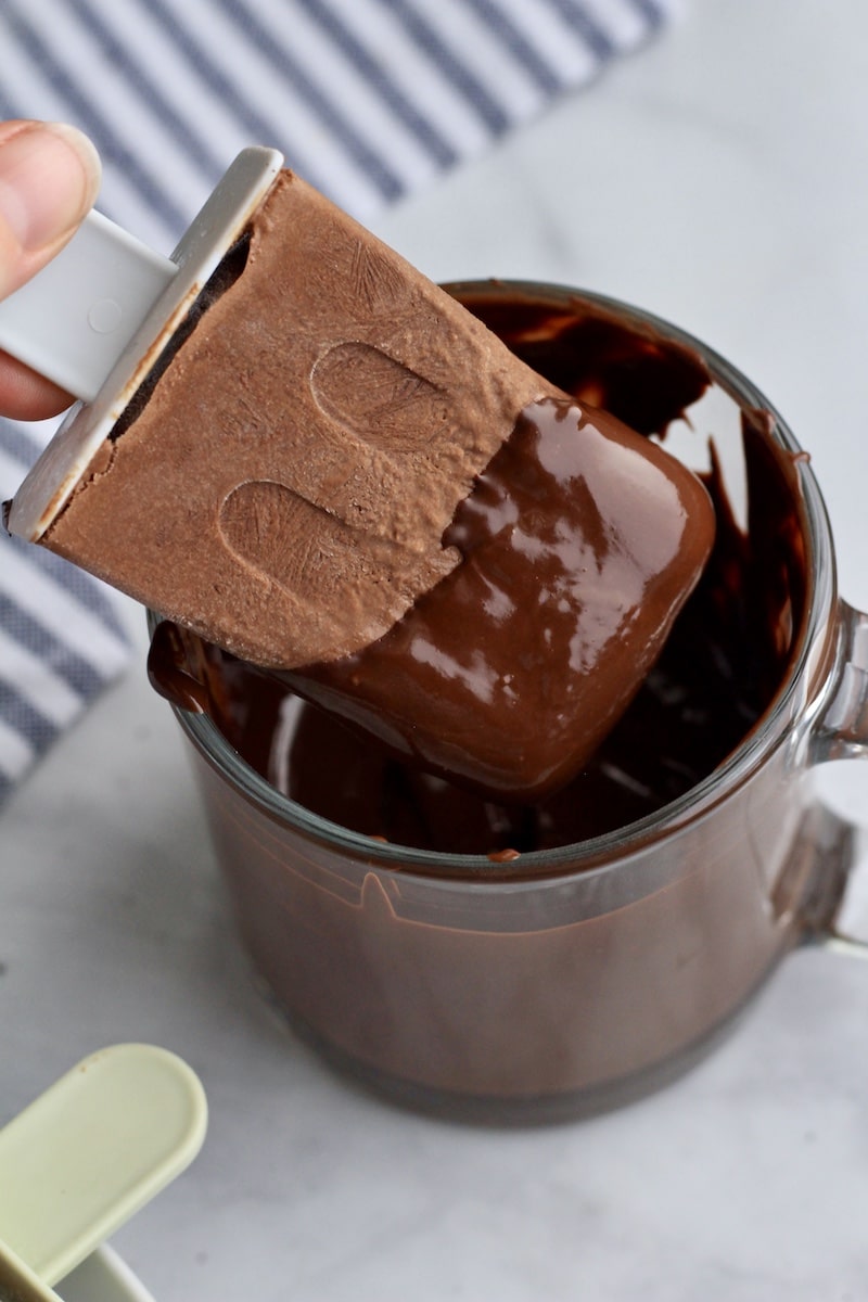 A hand dipping the vegan fudgesicle in a chocolate shell mixture.