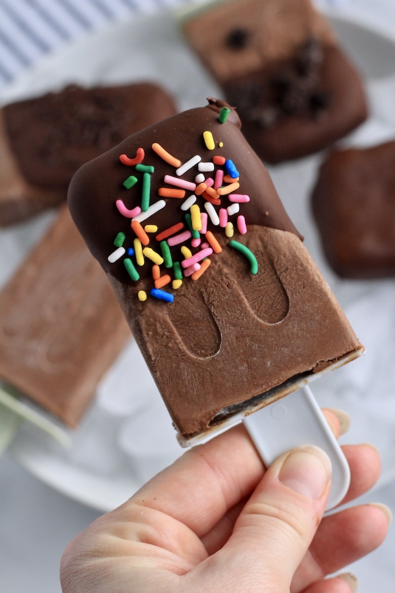 A hand holding a chocolate dipped vegan fudgesicle with rainbow sprinkles.