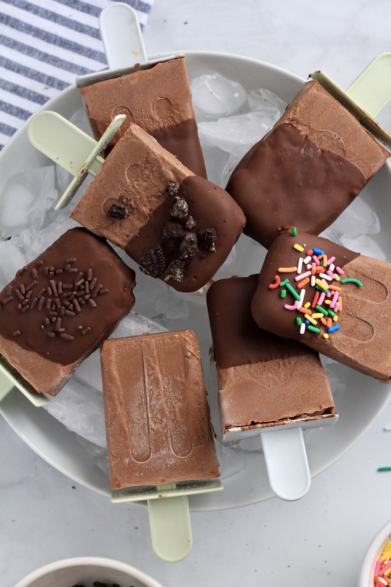 A bowl of vegan fudgesicles some dipped in chocolate some not, some sprinkled with sprinkles and Oreo crumbs.