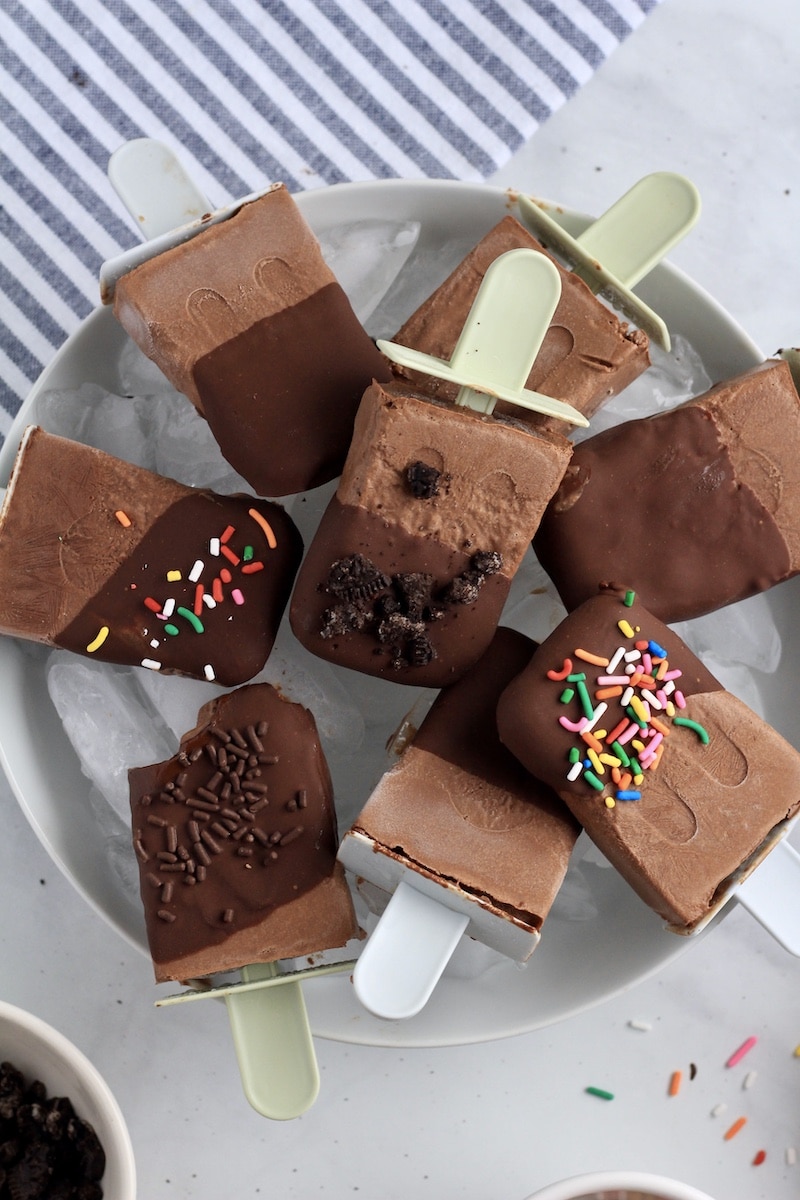 A bowl of vegan fudgesicles some dipped in chocolate some not, some sprinkled with sprinkles and Oreo crumbs and one missing a bite in the lower left section of the bowl.