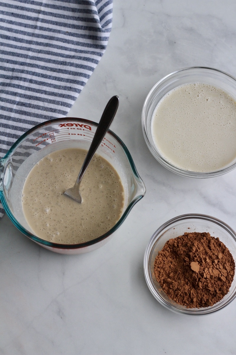 A bowl of oat milk next to a liquid measuring sup with coconut cream, sugar, and vanilla, next to a small bowl of cocoa powder.