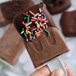 A hand holding a chocolate dipped and rainbow sprinkled vegan fudgesicle with a blue popsicle stick.