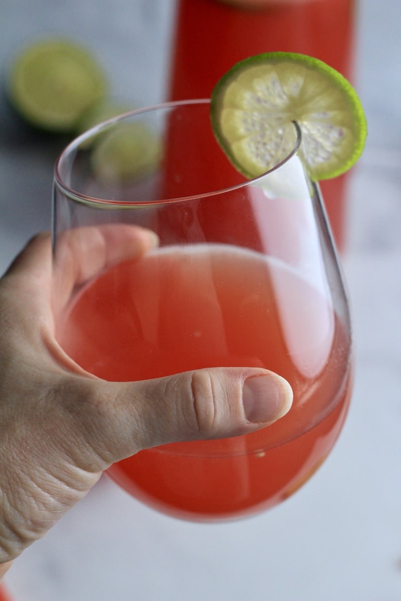 A hand holding a glass of watermelon limeade with a slice of lime on the glass.