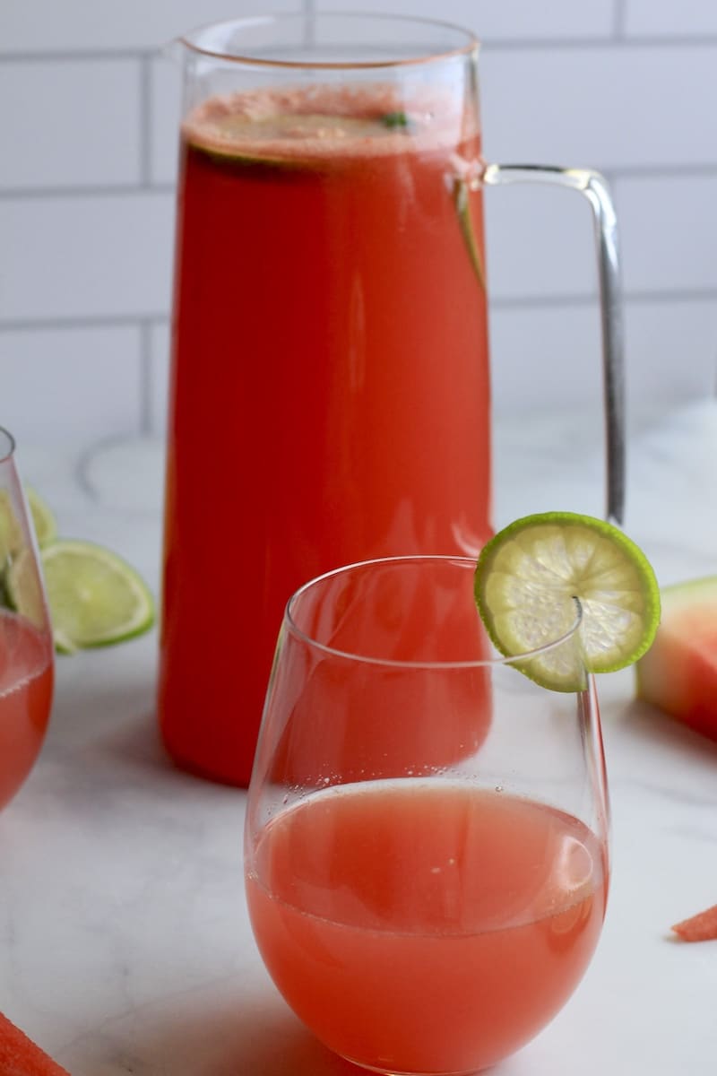 A cup of watermelon limeade with a slice of lime on the side with a pitcher of limeade in the back.