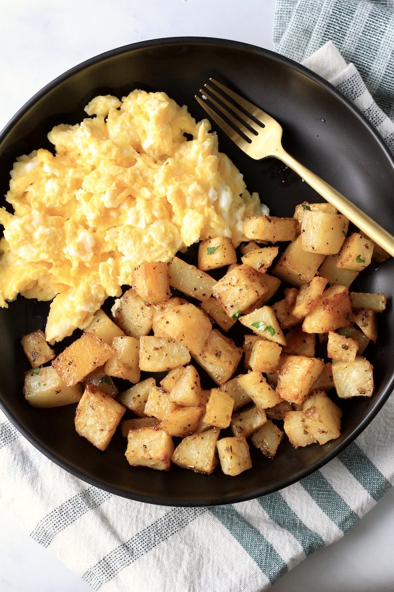 A black plate on a green and white dish towel with roasted potatoes, eggs, and a gold fork.