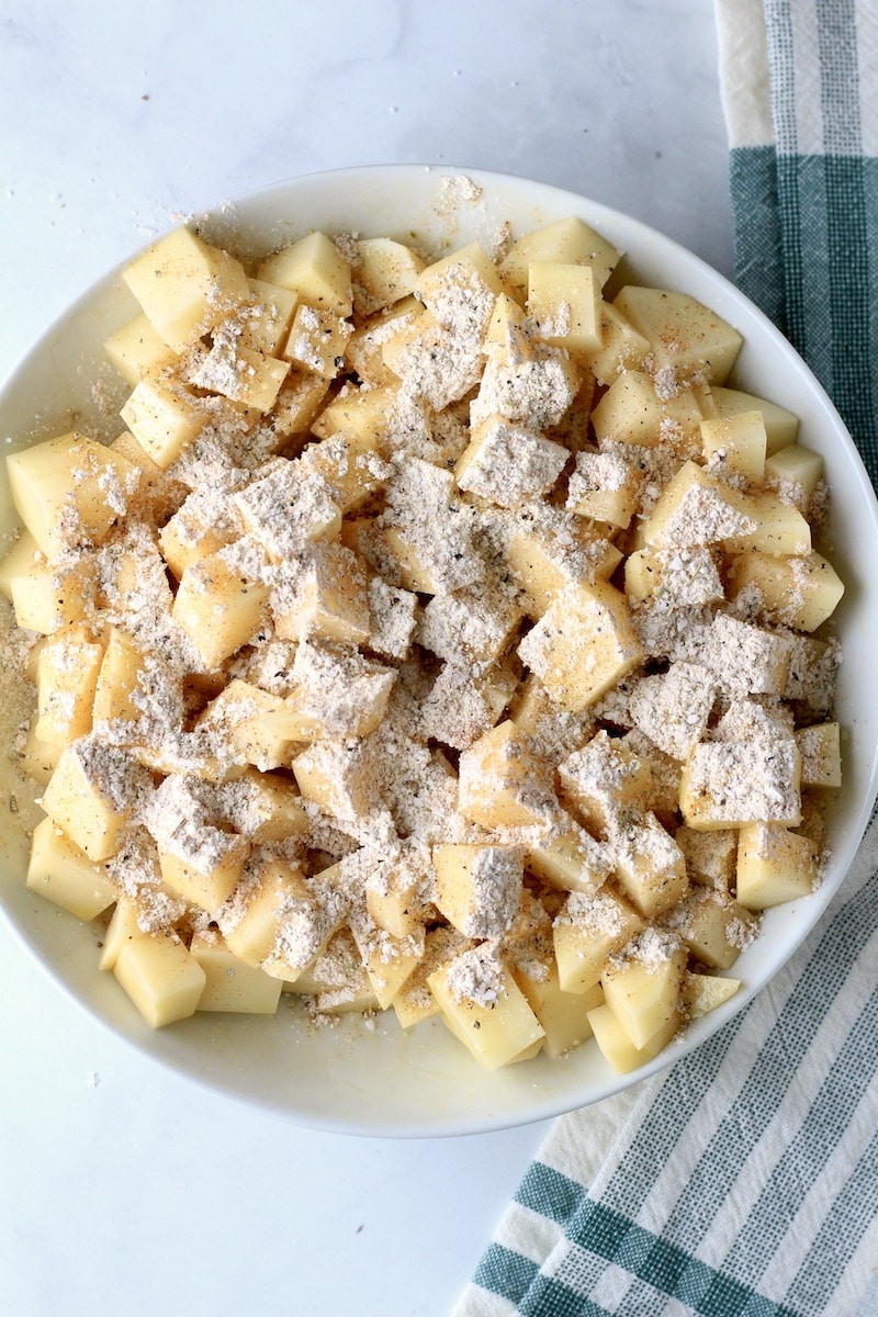 A bowl of diced potatoes topped with seasoning on a counter with a white and green dish towel to the right.