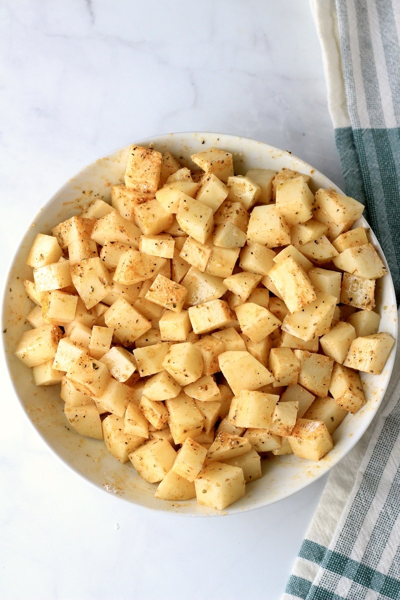 A white bowl with diced potatoes coated in oil and seasoning.