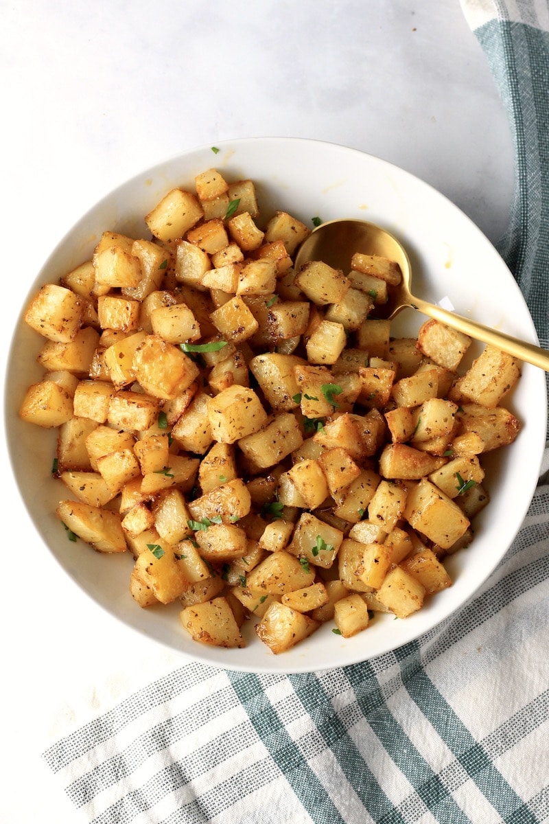A white bowl with roasted breakfast potatoes and a gold spoon on a counter with a green and white towel.