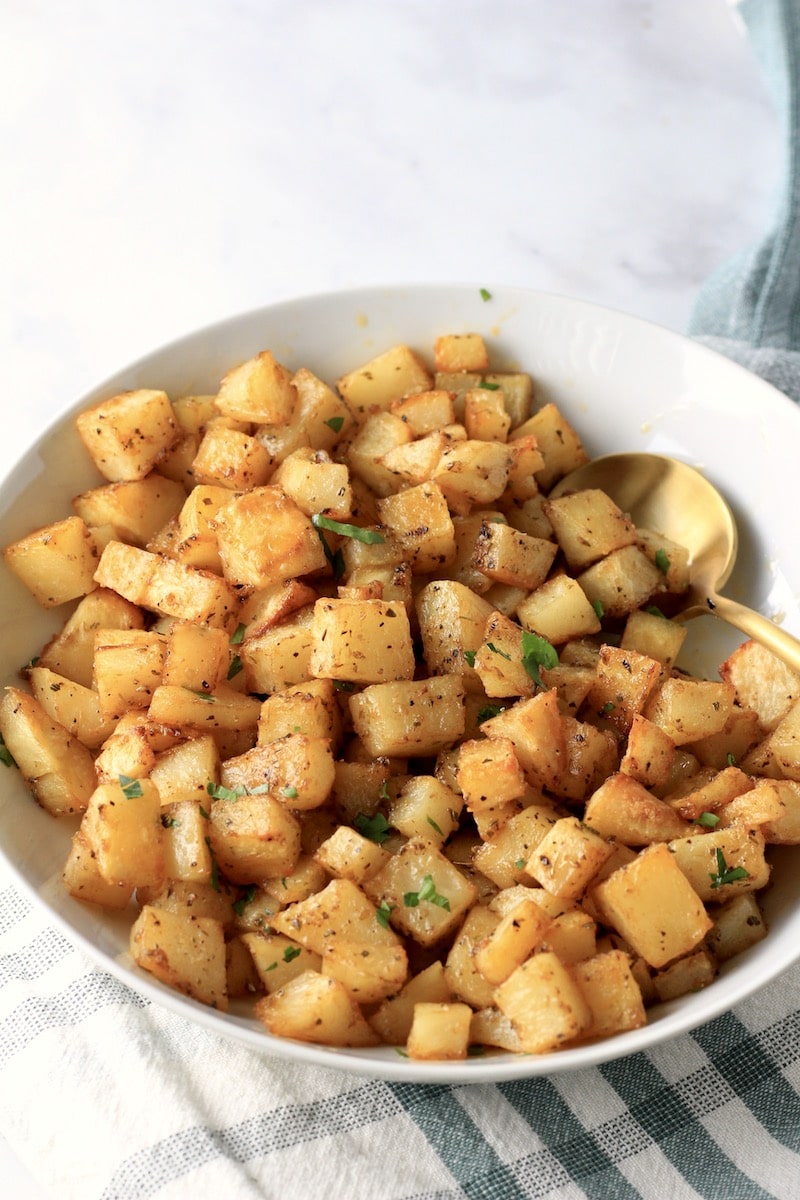 A bowl of oven-roasted breakfast potatoes with a gold spoon to the right and a green and white towel on the counter.