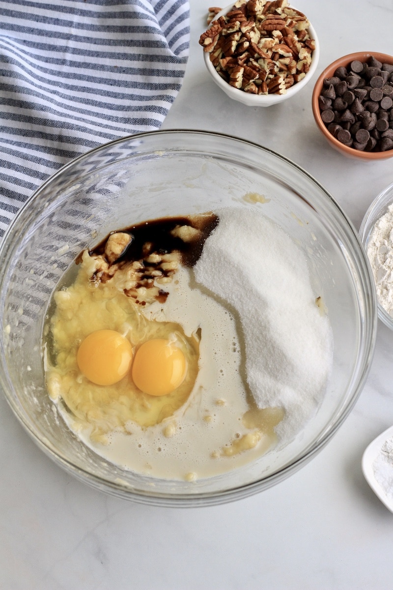 A large glass bowl with banana, sugar, eggs, and vanilla before mixing.