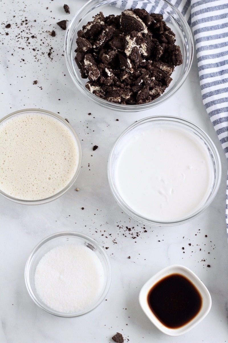 Ingredients for cookies and cream popsicles in bowls on a white counter.